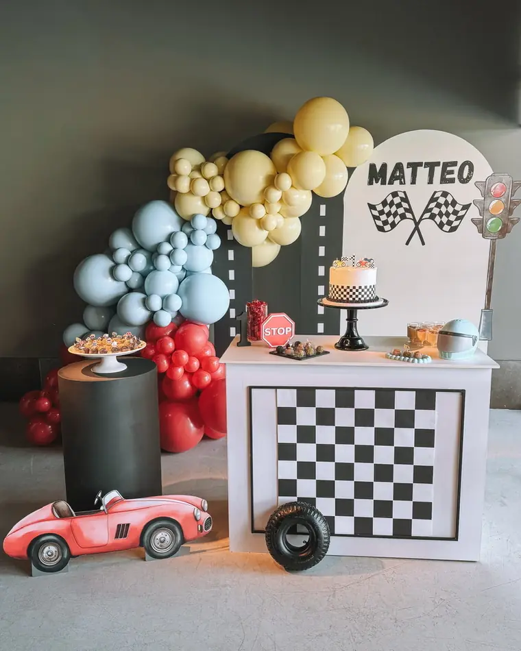 Mesa de festa com tema de carros de corrida: balões azuis e bege, painel axadrezado e um carro vermelho de brincar.
