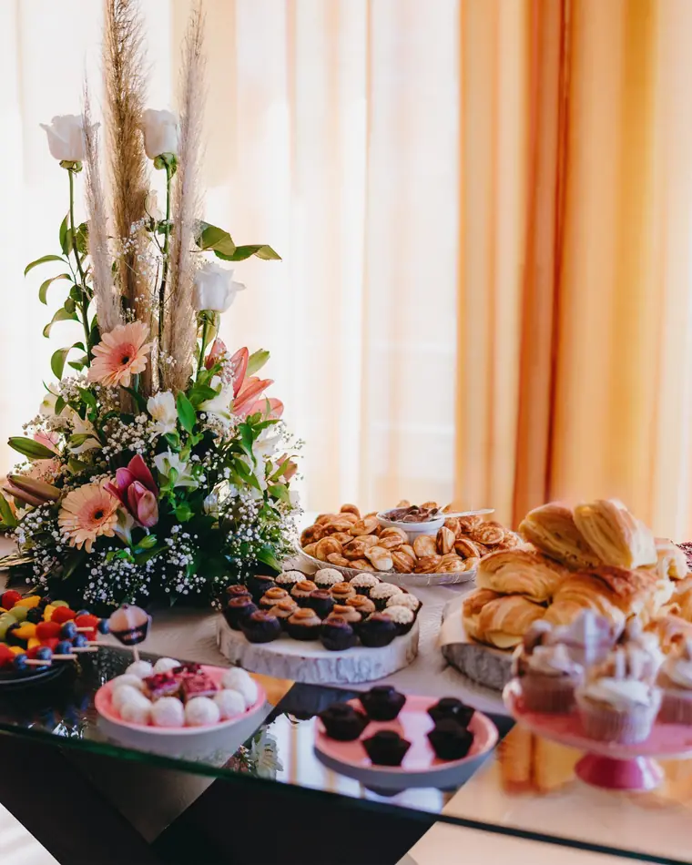 Mesa de doces e salgados com arranjo floral e cortinados cor de laranja ao fundo.