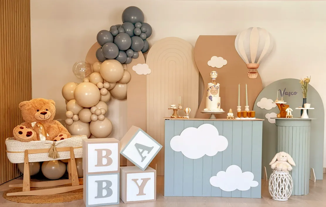 Cenário de baby shower com balões azuis e bege, ursinho de peluche, cubos com letras e um balão de ar quente decorativo.