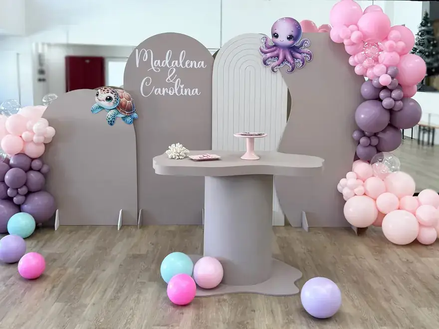 Cenário de festa em tons de lilás e roxo com balões e mesa de bolo.